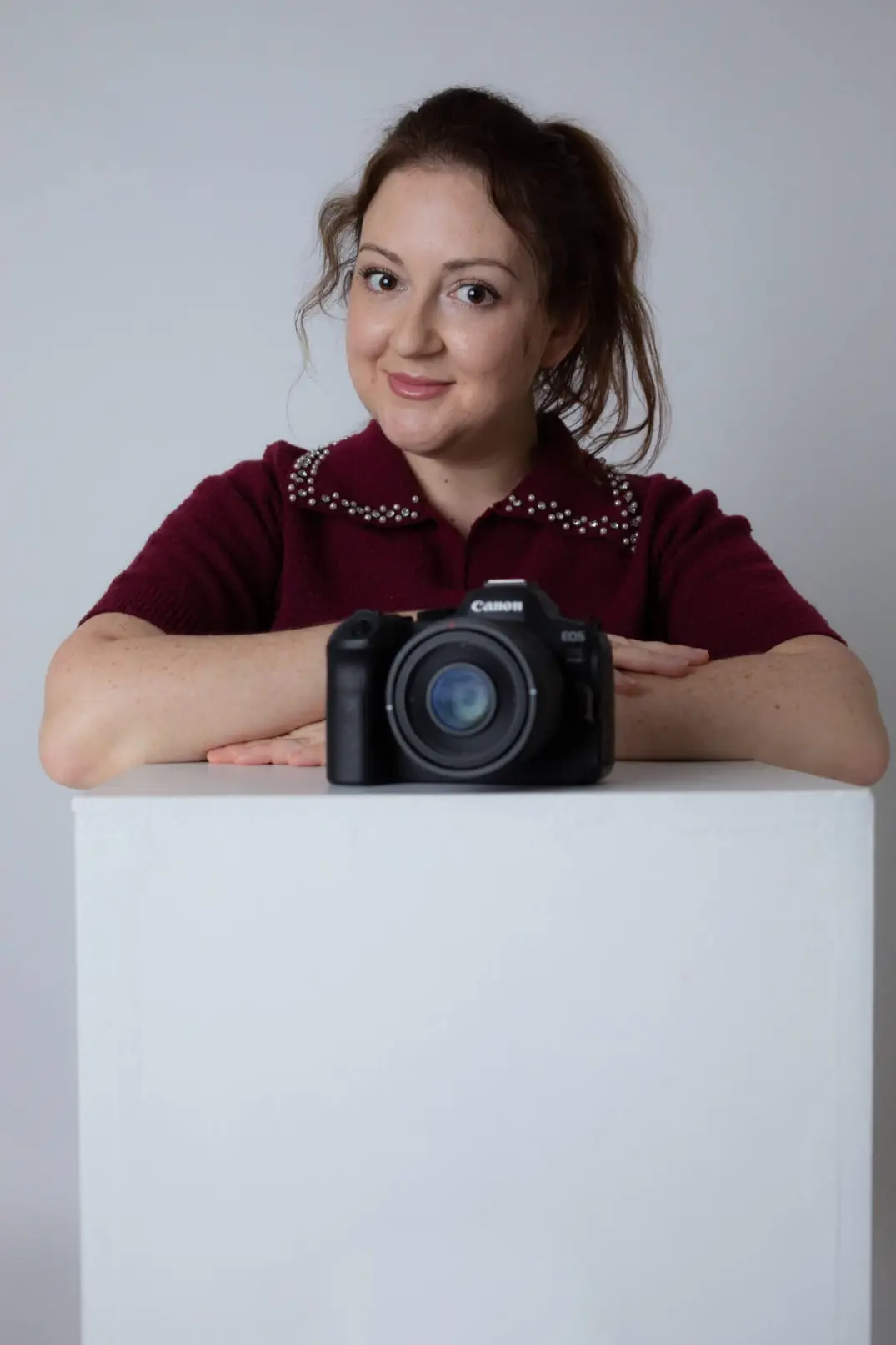 Natalia Vaxman leaning on a white pedestal with her professional camera in a clean studio setting.